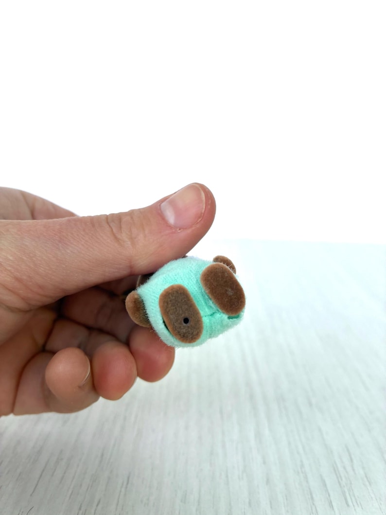 May include: A miniature plush panda bear toy in a light teal color with brown accents. The small toy has brown ears and feet. The panda is held in a person's hand against a white background.