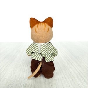 May include: A small, brown and tan cat figurine, viewed from the back. The cat wears a green and white striped shirt and brown pants. The cat's tail is tan. The figurine is on a light wood surface.