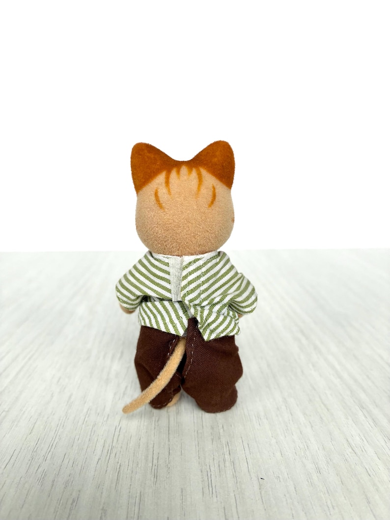 May include: A small, brown and tan cat figurine, viewed from the back. The cat wears a green and white striped shirt and brown pants. The cat's tail is tan. The figurine is on a light wood surface.