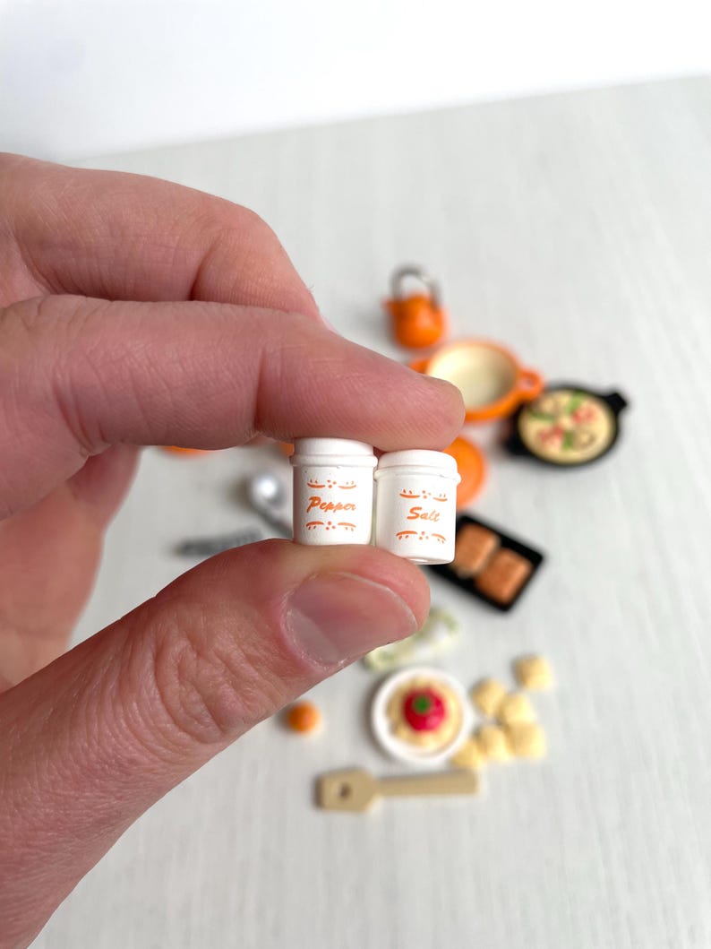 May include: Two miniature white salt and pepper shakers with orange lettering. The shakers say "Salt" and "Pepper" on them.