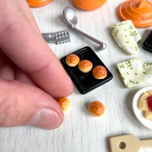 May include: A miniature kitchen set with a variety of items, including a small orange pot with a lid, a small black baking sheet with four small bread rolls, a small orange pot with a lid, a small wooden spoon, and a small white plate with a small pastry.