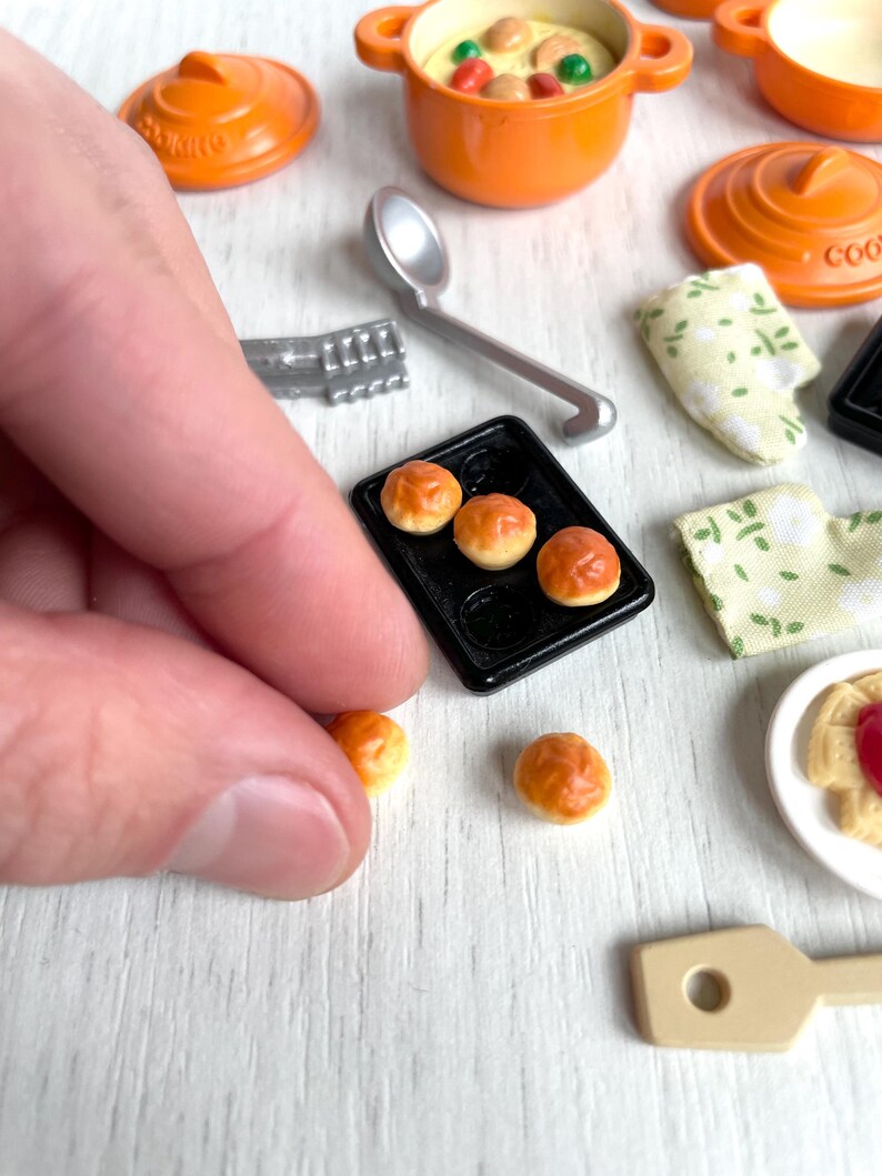 May include: A miniature kitchen set with a variety of items, including a small orange pot with a lid, a small black baking sheet with four small bread rolls, a small orange pot with a lid, a small wooden spoon, and a small white plate with a small pastry.