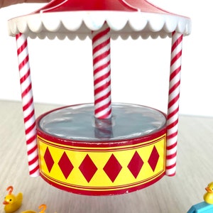Op de afbeelding: Een carnavalsspel met rode en witte strepen, een doorzichtig plastic vijvertje en gele rubberen eendjes. De vijver is omgeven door rode en witte gestreepte palen en een rood dak met een witte gegolfde rand.