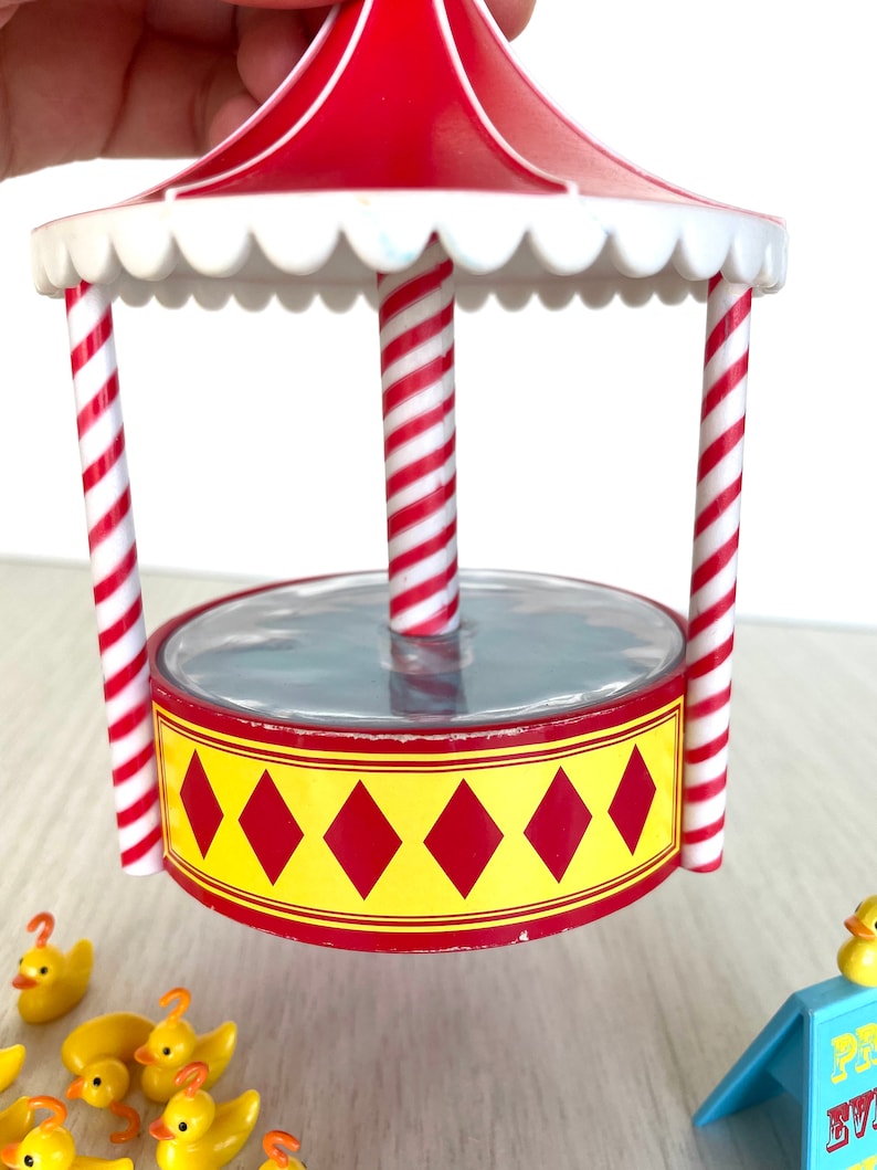 Op de afbeelding: Een carnavalsspel met rode en witte strepen, een doorzichtig plastic vijvertje en gele rubberen eendjes. De vijver is omgeven door rode en witte gestreepte palen en een rood dak met een witte gegolfde rand.
