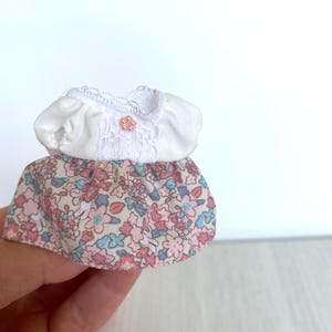 May include: A miniature white dress with a floral print skirt. The dress has a lace trim and a small pink button.