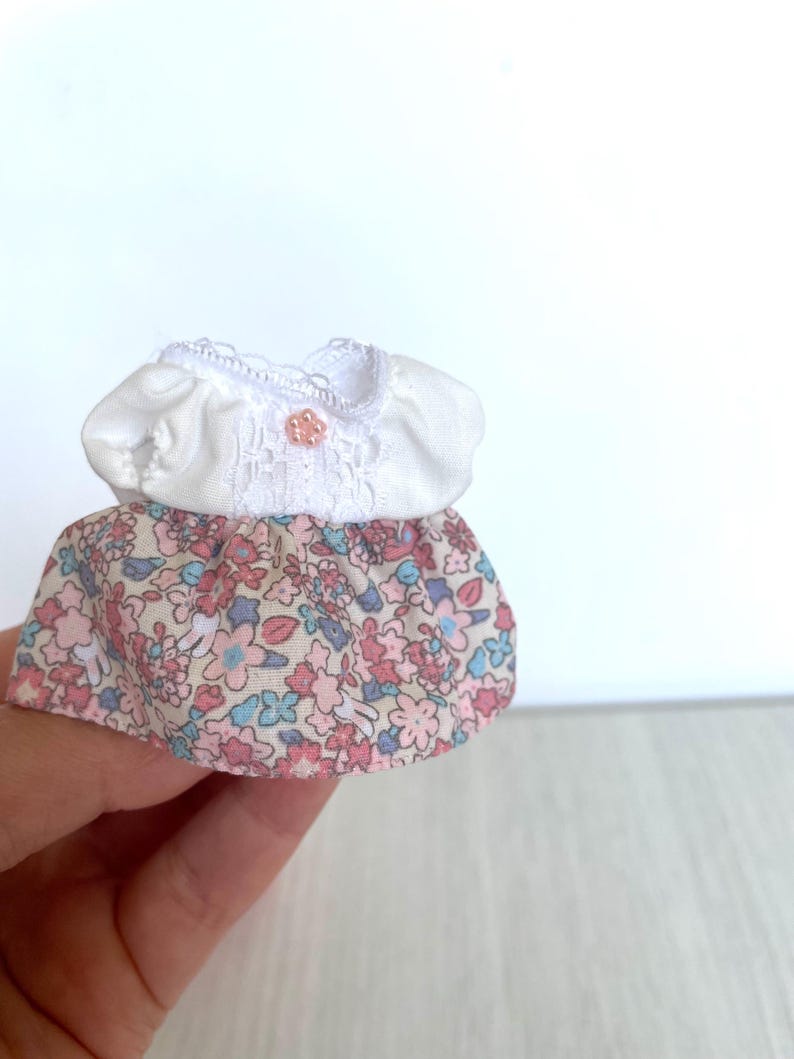 May include: A miniature white dress with a floral print skirt. The dress has a lace trim and a small pink button.