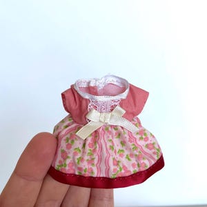 May include: A pink and white floral print dress with a white lace trim and a white satin bow. The dress has a red ribbon trim around the bottom.