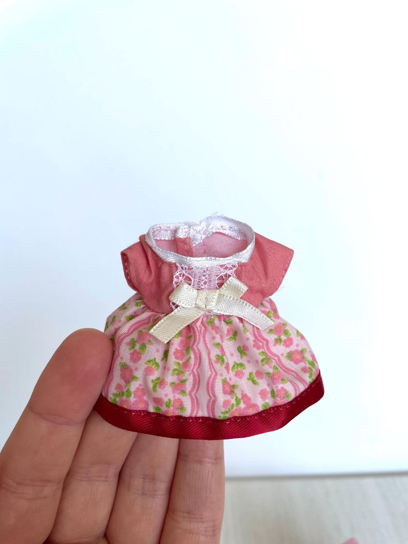 May include: A pink and white floral print dress with a white lace trim and a white satin bow. The dress has a red ribbon trim around the bottom.