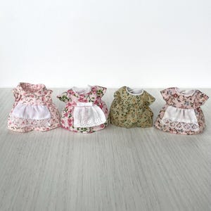 May include: Four miniature floral print dresses with white lace trim and aprons. The dresses are pink, green, and two shades of pink.
