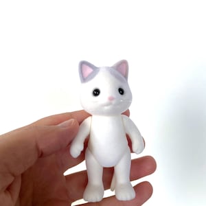 May include: A white cat figurine with gray ears and a pink nose. The cat has black eyes and is standing on a white background.