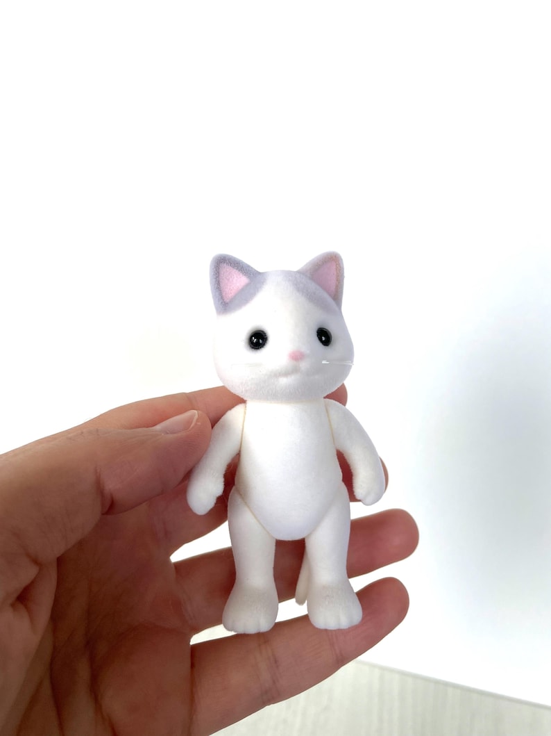May include: A white cat figurine with gray ears and a pink nose. The cat has black eyes and is standing on a white background.