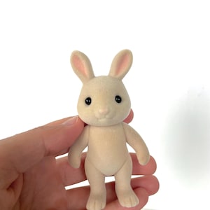 May include: A small, cream-colored rabbit figurine with pink ears and black eyes. The rabbit is standing upright with its feet together.