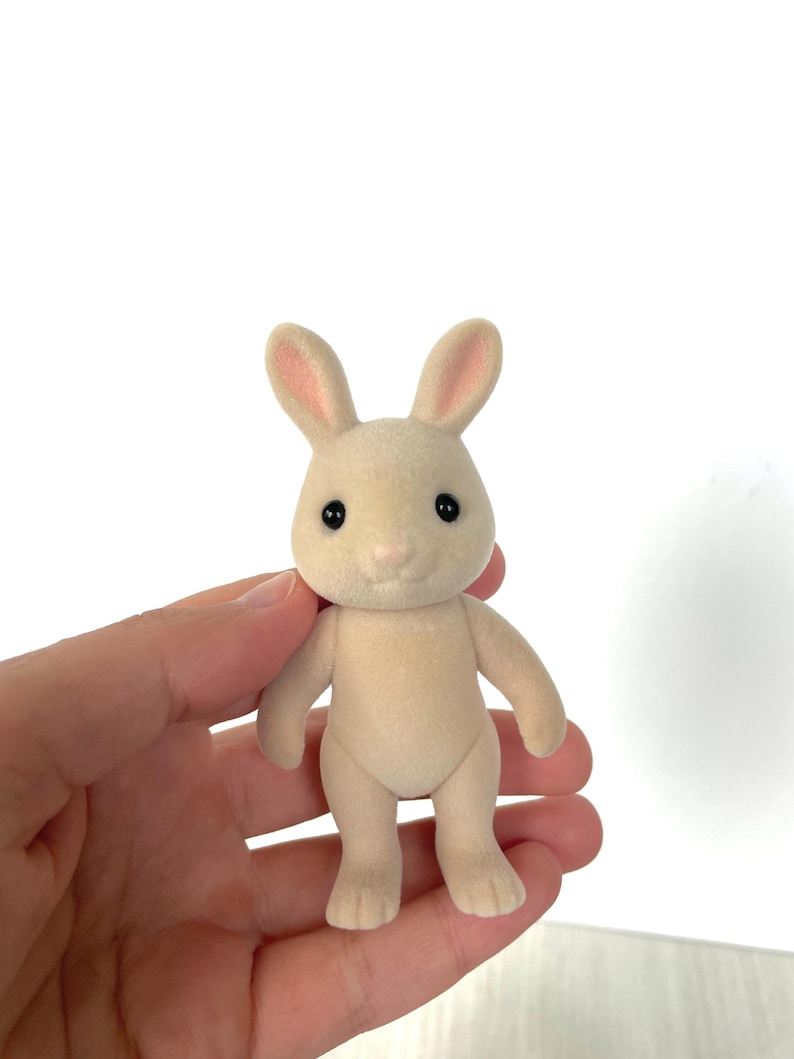 May include: A small, cream-colored rabbit figurine with pink ears and black eyes. The rabbit is standing upright with its feet together.