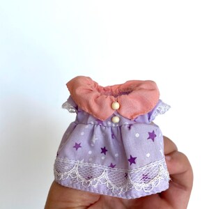 May include: A purple and pink doll dress with white polka dots and purple stars. The dress has a lace trim and a pink collar with a white button.