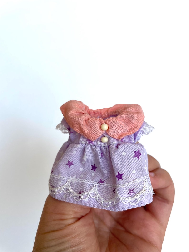 May include: A purple and pink doll dress with white polka dots and purple stars. The dress has a lace trim and a pink collar with a white button.