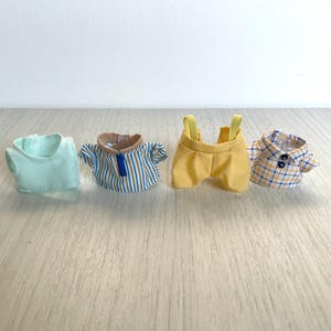 Puede incluir: Cuatro prendas de vestir en miniatura para una muñeca o una figura de acción. Los artículos incluyen una camisa sin mangas azul claro, una camisa a rayas azul y blanca, un mono amarillo y una camisa a cuadros azules y amarillos.