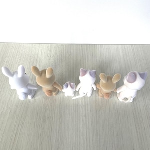 May include: A group of six plush animal figurines, including a white rabbit, a brown rabbit, a white cat, a brown cat, and two smaller white cats. The figurines are arranged in a line on a light wood surface.