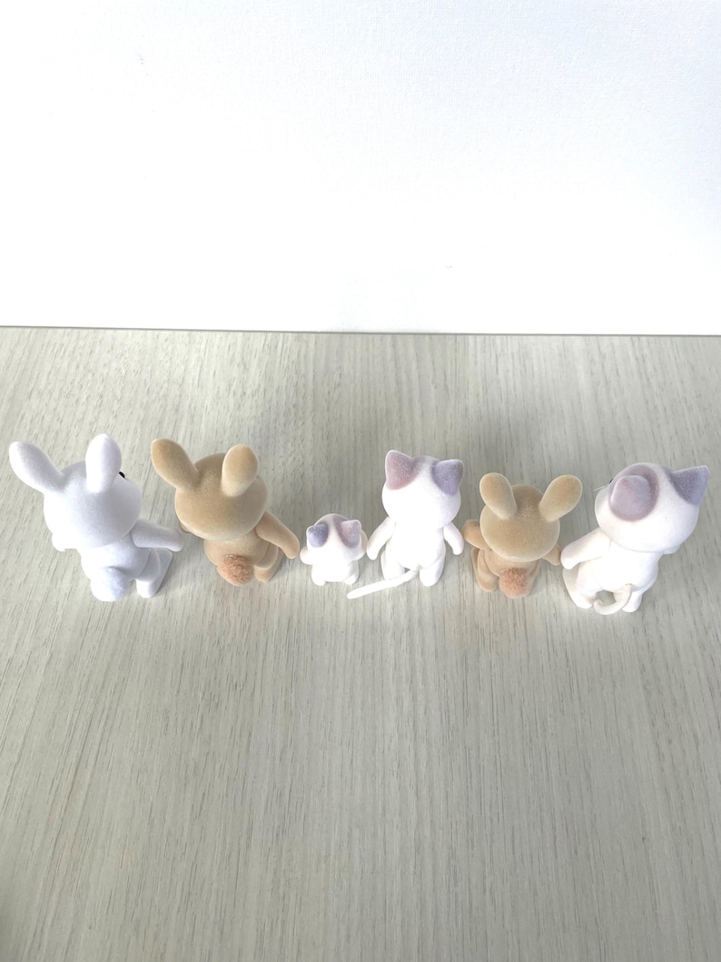 May include: A group of six plush animal figurines, including a white rabbit, a brown rabbit, a white cat, a brown cat, and two smaller white cats. The figurines are arranged in a line on a light wood surface.