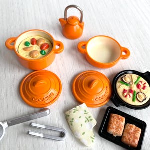May include: A set of miniature orange cookware, including a teapot, two pots with lids, a frying pan, and two baking dishes. The cookware is decorated with the word "Cooking" and features a variety of food items, including a pizza, bread, and a casserole.
