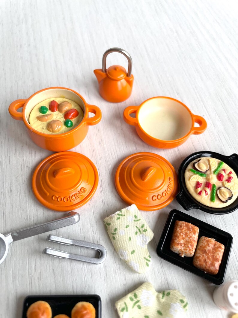May include: A set of miniature orange cookware, including a teapot, two pots with lids, a frying pan, and two baking dishes. The cookware is decorated with the word "Cooking" and features a variety of food items, including a pizza, bread, and a casserole.