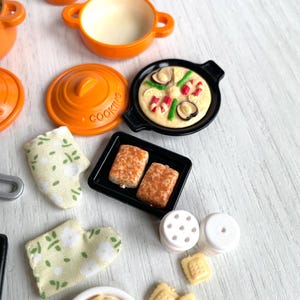 May include: A set of miniature kitchen cookware and food items, including a teapot, pots, pans, a baking sheet, salt and pepper shakers, and cookies. The items are orange, black, and white.