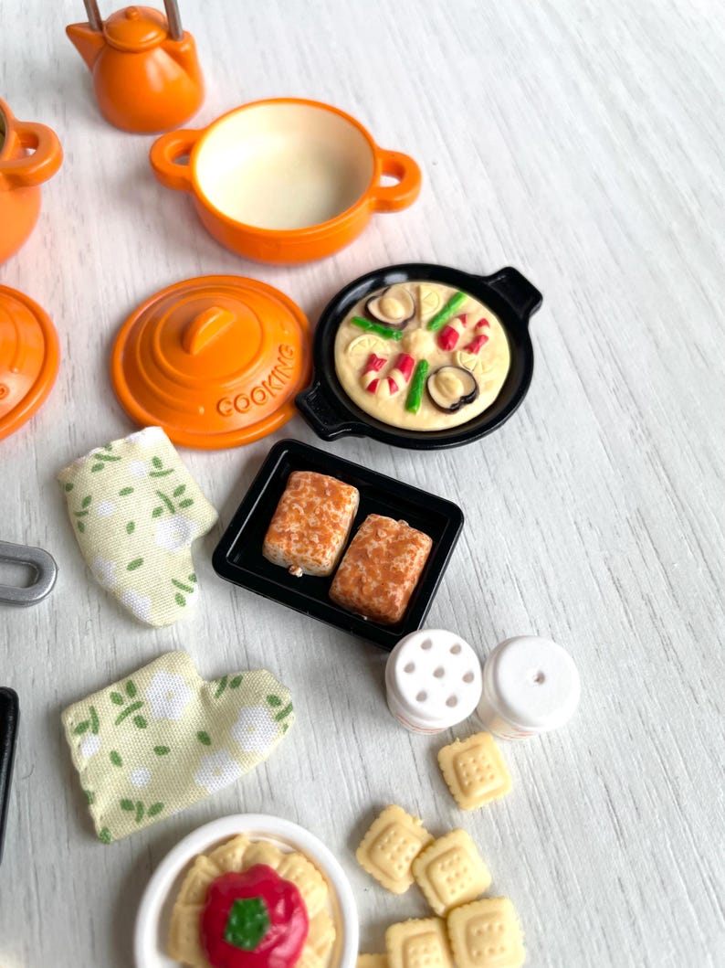 May include: A set of miniature kitchen cookware and food items, including a teapot, pots, pans, a baking sheet, salt and pepper shakers, and cookies. The items are orange, black, and white.