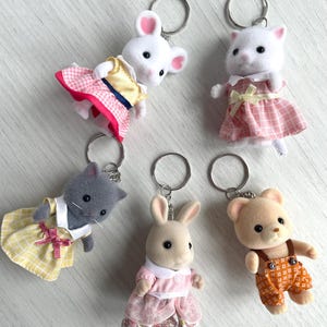 Op de afbeelding: Vijf pluche dieren sleutelhangers met zilveren sleutelringen. De dieren zijn een witte muis met een roze en gele jurk, een witte kat met een roze en gele jurk, een grijze kat met een gele jurk, een bruin konijn met een roze jurk en een bruine beer met een oranje overall.