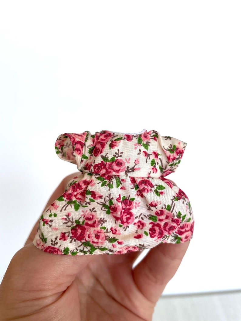May include: A small, white dress with a pink and green floral print. The dress has a ruffled neckline and short sleeves.