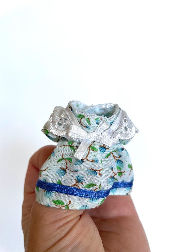 May include: A miniature blue and white floral dress with a white lace collar and blue ribbon bow. The dress has a blue ribbon trim around the bottom.