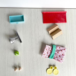 Może przedstawiać: Zestaw miniaturowych mebli do domku dla lalek, w tym czerwony dach, niebieska szafka, drewniane schody, zielona konewka, srebrny dzbanek, para białych i czerwonych solniczek i pieprzniczek, para żółtych talerzy i różowo-zielony materiał w kwiatowy wzór.