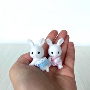 May include: Two white plush bunny figurines wearing blue and pink outfits. The bunnies are sitting in the palm of a hand.