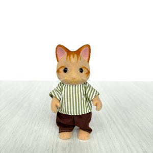 May include: A small, tan cat figurine with orange stripes on its head and ears. It wears a green and white striped shirt and brown pants. The figurine has a friendly expression and is standing upright.