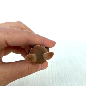 May include: A small, brown, flocked toy animal with large ears and a dark nose and eyes. The toy is held in a person's hand against a white background. The toy is a miniature figurine.