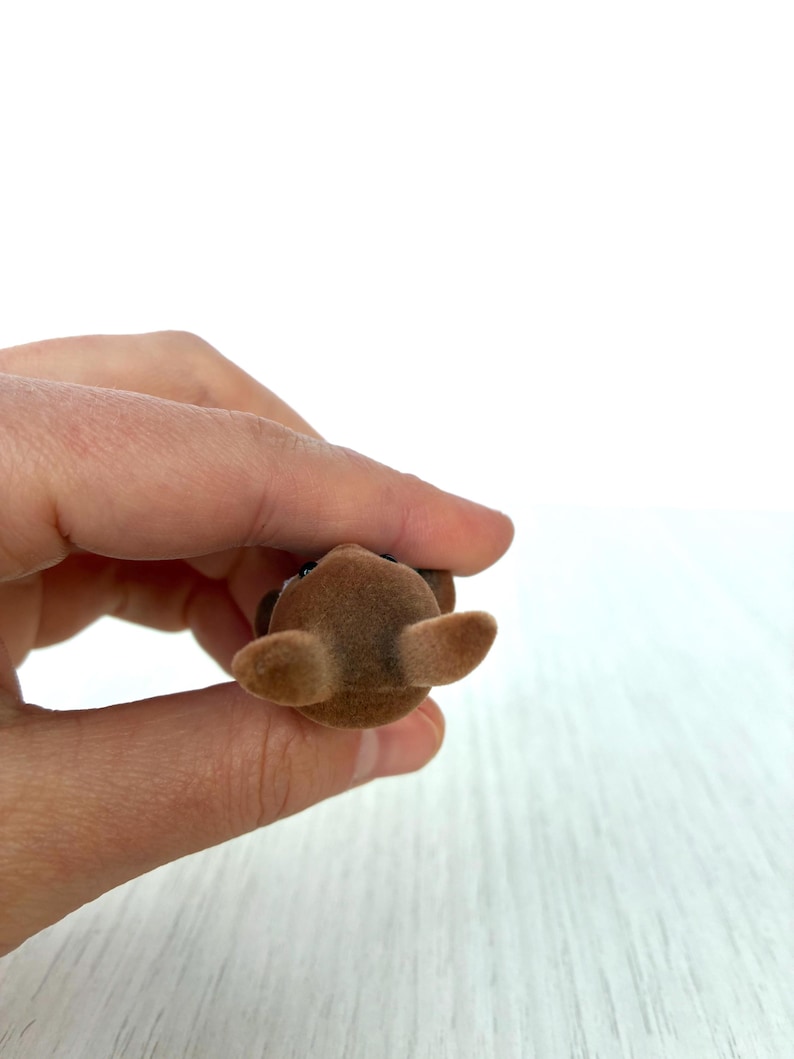 May include: A small, brown, flocked toy animal with large ears and a dark nose and eyes. The toy is held in a person's hand against a white background. The toy is a miniature figurine.