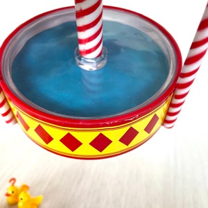 Op de afbeelding: Een rood-wit gestreepte carrouselpaal met een blauw waterbekken en een geel ruitpatroon rond de rand. De carrousel hangt boven een houten oppervlak met gele rubberen eendjes.