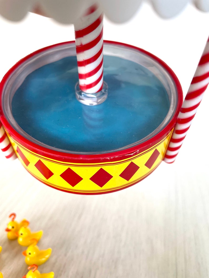 Op de afbeelding: Een rood-wit gestreepte carrouselpaal met een blauw waterbekken en een geel ruitpatroon rond de rand. De carrousel hangt boven een houten oppervlak met gele rubberen eendjes.