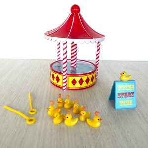 Op de afbeelding: Een rood-wit gestreept kermisspel met een gele basis en een blauw bord met de tekst "Prijs elke keer". Er zijn negen gele rubberen eendjes en twee gele stokken met ringen aan de uiteinden.