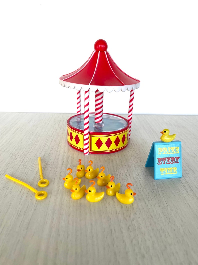 Op de afbeelding: Een rood-wit gestreept kermisspel met een gele basis en een blauw bord met de tekst "Prijs elke keer". Er zijn negen gele rubberen eendjes en twee gele stokken met ringen aan de uiteinden.