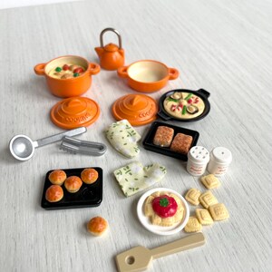 May include: A miniature kitchen playset with orange pots, pans, and a teapot. There are also miniature food items, including pizza, bread, and pastries. The set includes a spatula, tongs, and two salt and pepper shakers.