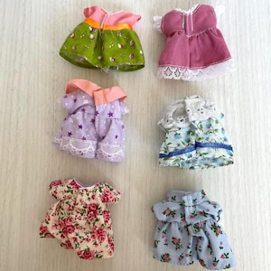 May include: Six miniature doll dresses in various floral patterns and colors, including green, pink, purple, blue, and white. The dresses have lace trim and some have buttons.