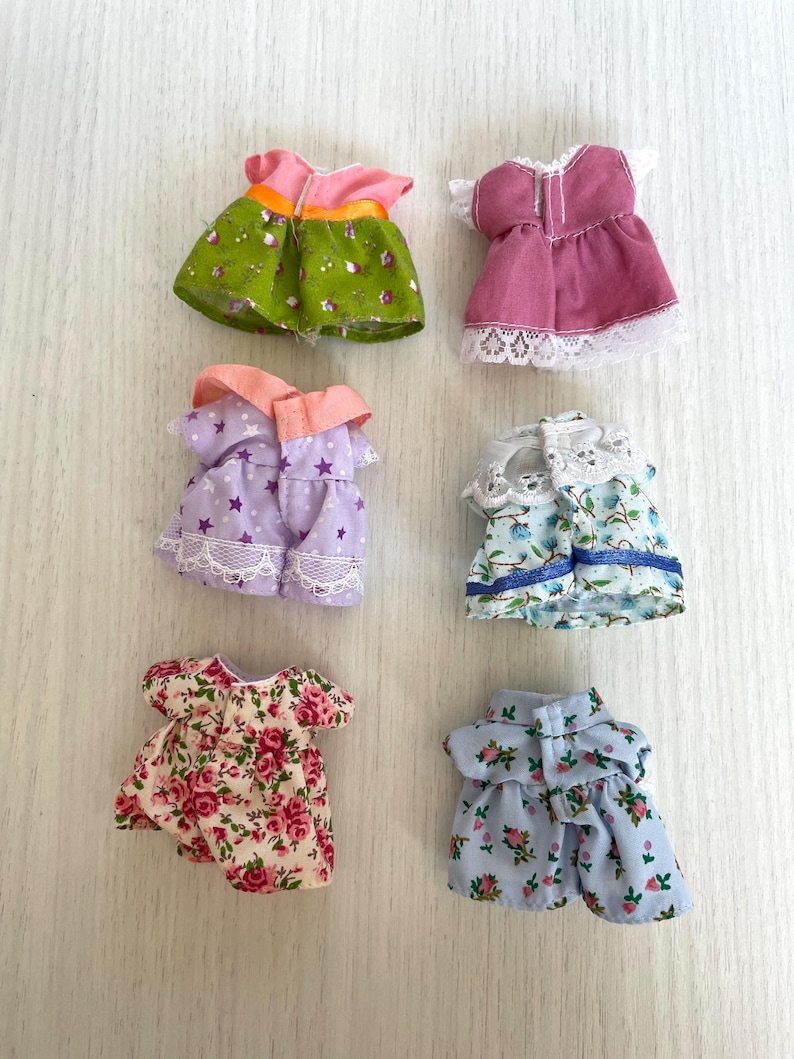 May include: Six miniature doll dresses in various floral patterns and colors, including green, pink, purple, blue, and white. The dresses have lace trim and some have buttons.