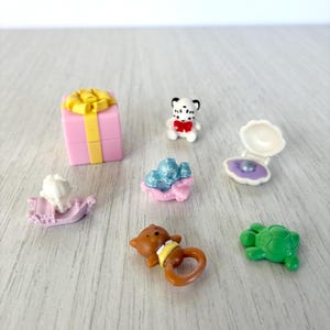 Peut inclure: Ensemble de jouets miniatures comprenant une boîte cadeau rose avec un nœud jaune, un ours blanc et noir avec un nœud rouge, un coquillage avec une perle violette, un cheval à bascule, un service à thé bleu et rose, un ours brun avec un anneau jaune et une tortue verte.