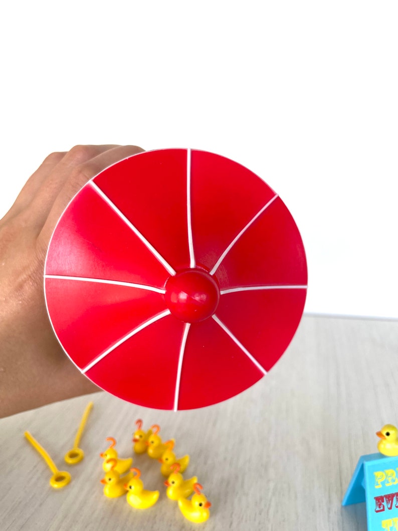 Op de afbeelding: Een rode en witte plastic speelgoedparaplu met een geel rubberen eendjespel. De eendjes staan in een rij op een houten oppervlak.