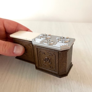 Könnte beinhalten: Ein Miniatur-Küchenherd in Braun mit einer weißen Arbeitsplatte und silbernen Brennern. Der Herd hat dekorative Rosendetails an den Schranktüren. Eine Hand hält den Miniaturherd.