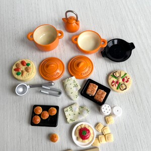 May include: A set of miniature kitchen cookware and food items, including orange pots, pans, a kettle, a black skillet, and various food items like pizza, bread, and pasta. The items are arranged on a light wood surface.
