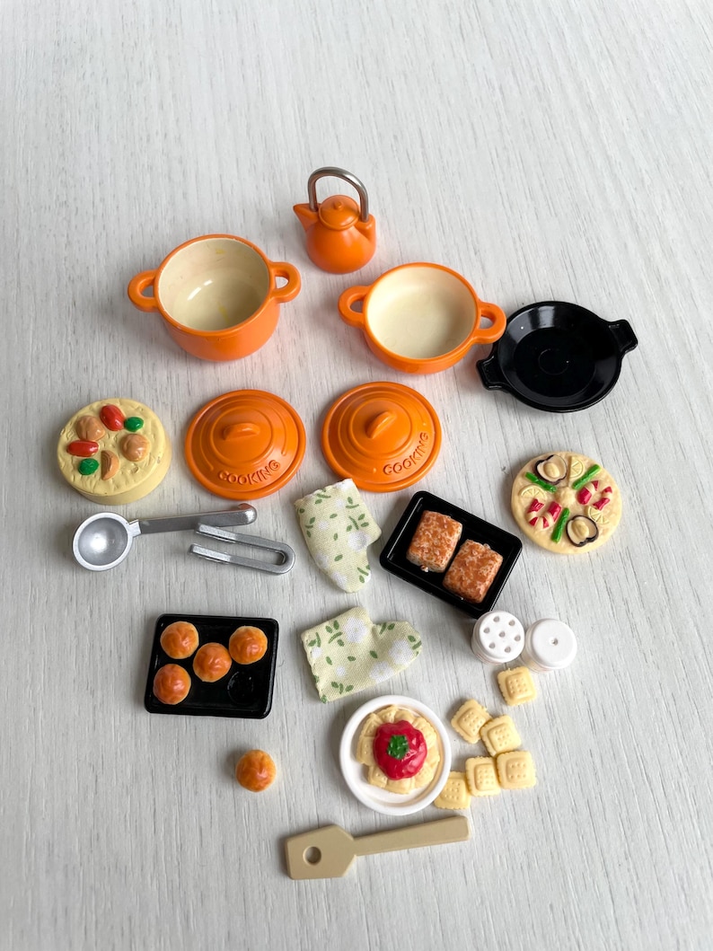 May include: A set of miniature kitchen cookware and food items, including orange pots, pans, a kettle, a black skillet, and various food items like pizza, bread, and pasta. The items are arranged on a light wood surface.