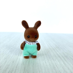 May include: A brown rabbit figurine wearing a mint green romper with a white lace collar. The toy rabbit has dark eyes and is standing upright. The figurine is a collectible toy.