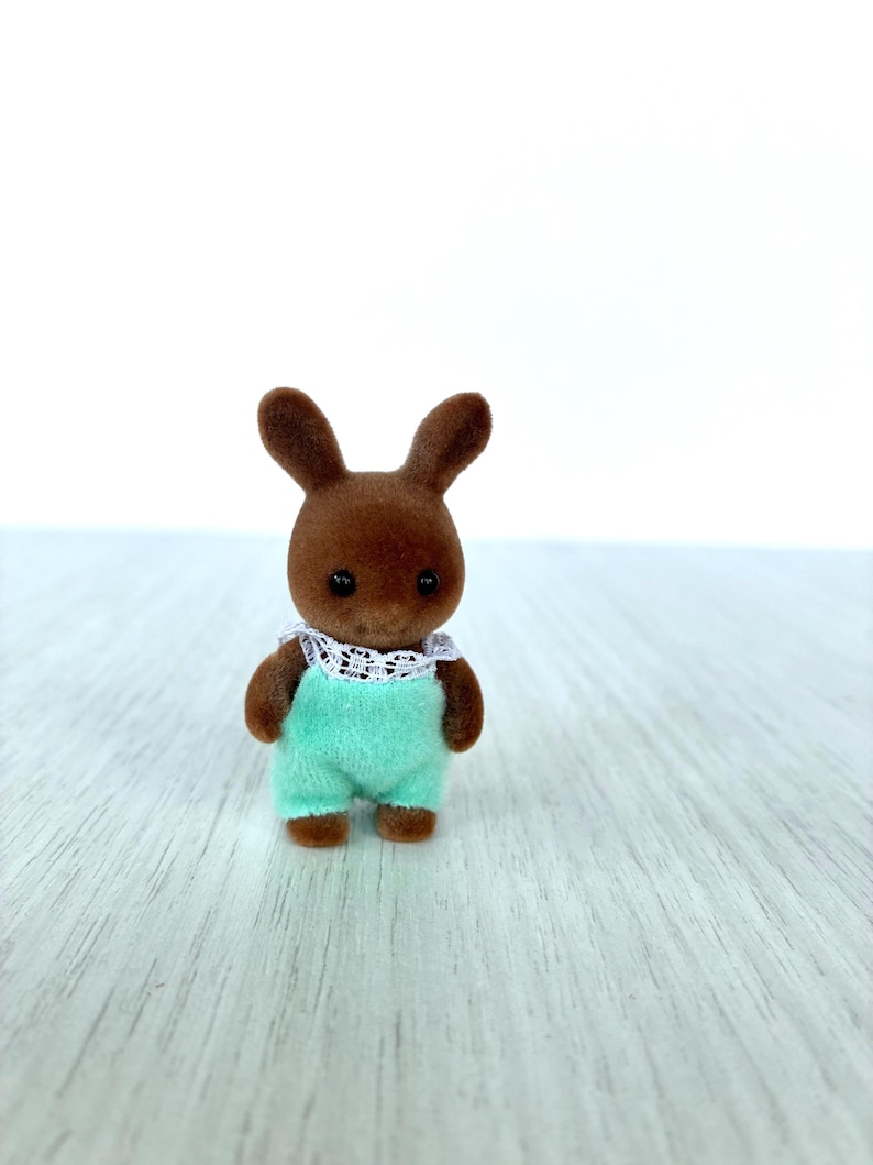May include: A brown rabbit figurine wearing a mint green romper with a white lace collar. The toy rabbit has dark eyes and is standing upright. The figurine is a collectible toy.