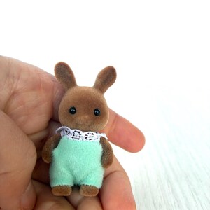 May include: A small brown rabbit figurine wearing a light green jumpsuit with a white lace collar. The toy has black eyes and small, upright ears. The figurine is held in a person's hand against a white background.