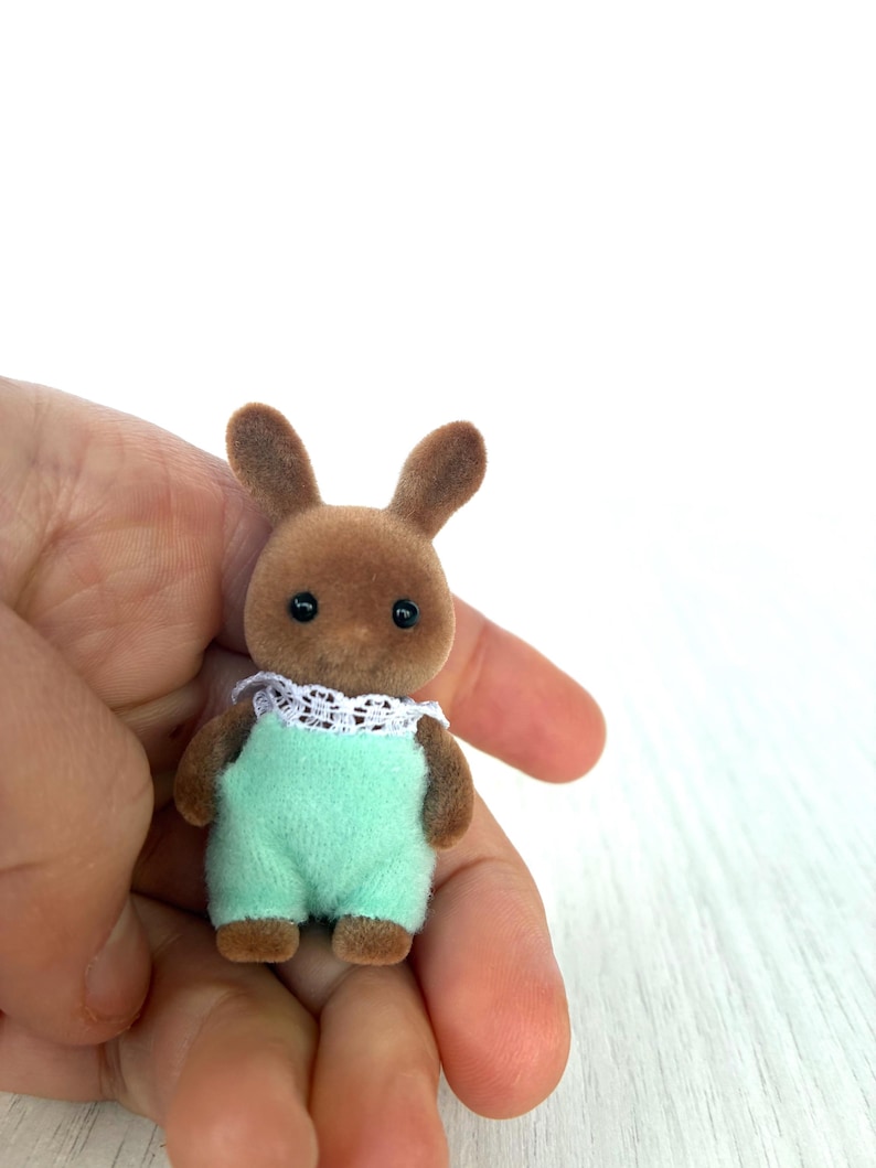 May include: A small brown rabbit figurine wearing a light green jumpsuit with a white lace collar. The toy has black eyes and small, upright ears. The figurine is held in a person's hand against a white background.